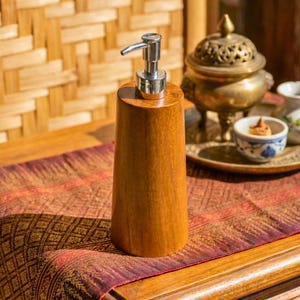 May include: A wooden soap dispenser with a silver pump. The dispenser is a light brown color and has a conical shape. It is sitting on a patterned red and gold fabric.