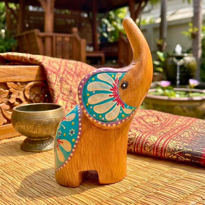 May include: A wooden elephant figurine with a raised trunk and painted details. The elephant is light brown with teal, red, and white floral patterns on its ears and body. The figurine is on a woven mat.