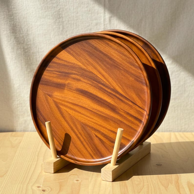 May include: A stack of three round wooden serving trays with a rich, warm brown grain pattern. The trays are displayed on a light-colored wooden stand. The trays are approximately 12 inches in diameter, perfect for serving food or displaying items.