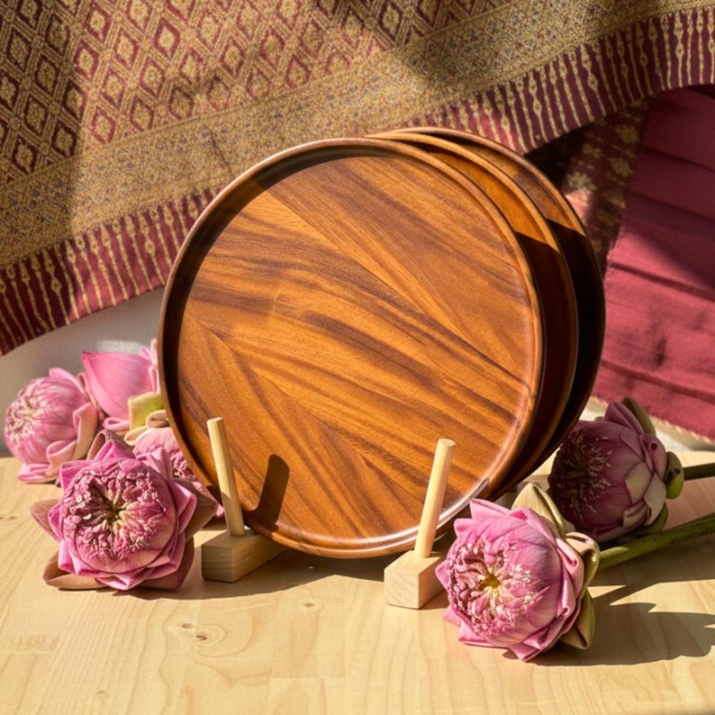 May include: A stack of three round wooden trays with a rich, warm wood grain. The trays are displayed on a light-colored wooden surface, accompanied by pink lotus flowers and small wooden stands. A patterned textile is in the background.
