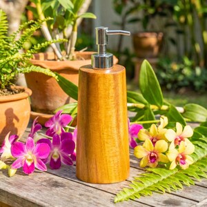 May include: A wooden soap dispenser with a silver pump. The dispenser is cylindrical and made of light brown wood. The background includes potted plants and colorful flowers, creating a natural, spa-like aesthetic.
