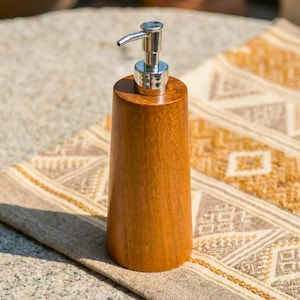 May include: A wooden soap dispenser with a silver pump. The dispenser is made of light brown wood and has a cylindrical shape that tapers slightly towards the base. The pump is made of shiny silver metal.