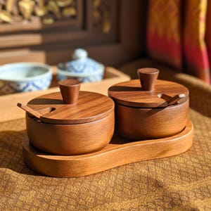 Può includere: Due barattoli di spezie in legno con coperchi e cucchiaini, posti su un vassoio in legno abbinato. I barattoli e il vassoio sono di colore marrone caldo, con venature naturali del legno visibili. I coperchi hanno piccole maniglie coniche.
