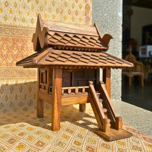 May include: A detailed miniature wooden house with a layered roof and a small staircase. The house is made of light brown wood and features intricate carvings. The design is reminiscent of traditional architecture.