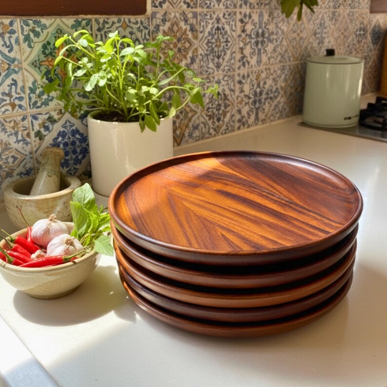 May include: A stack of six round wooden plates with a rich, warm brown color and visible wood grain. The plates are stacked on a white countertop, suggesting a kitchen or dining setting. The plates are approximately 10 inches in diameter.