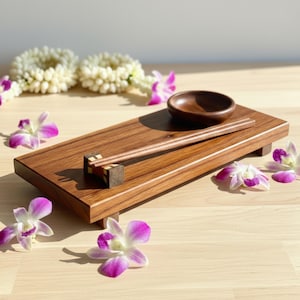 May include: A wooden sushi serving set featuring a rectangular tray, small bowl, and chopsticks. The tray is brown with a natural wood grain. The chopsticks rest on a decorative chopstick rest. Orchids in purple and white are scattered around.