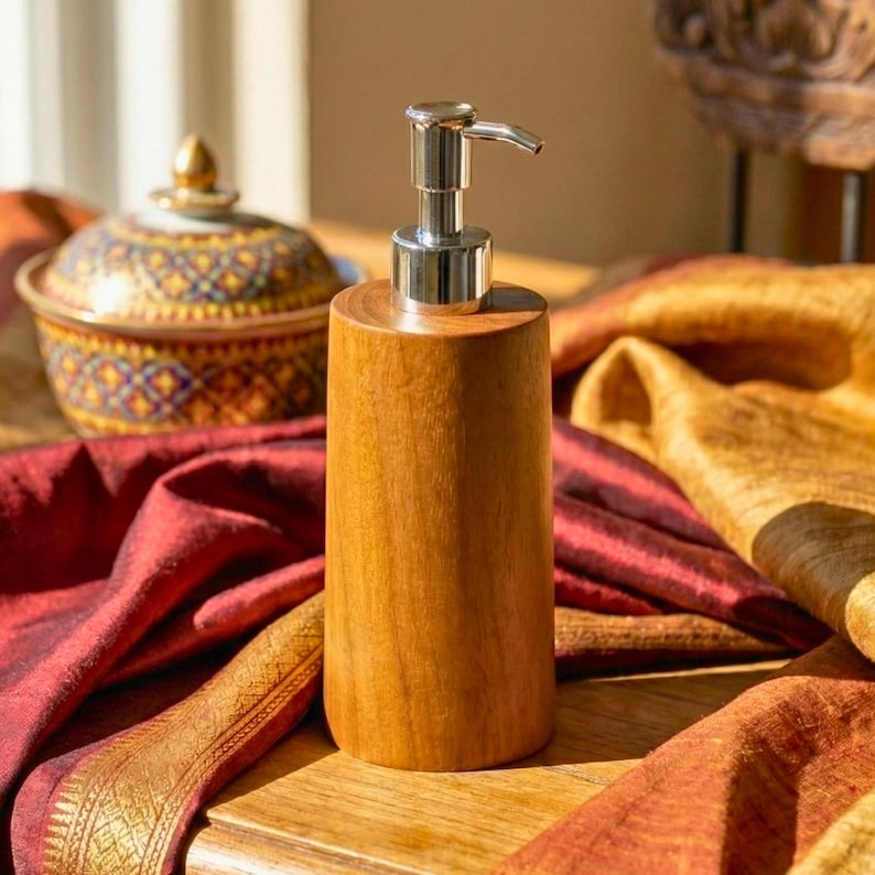 May include: A wooden soap dispenser with a silver pump. The dispenser is cylindrical and made of light brown wood. The background includes a decorative bowl and colorful fabric.