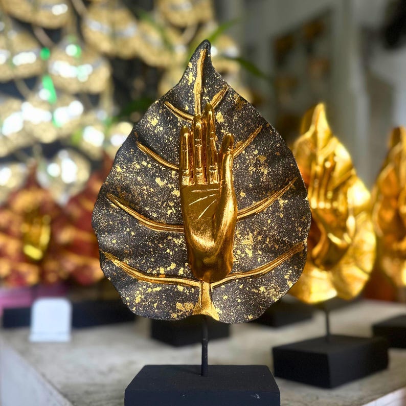 Pu&ograve; includere: Una scultura a forma di mano dorata che emerge da una forma a foglia con una finitura maculata nera e oro. La scultura &egrave; montata su una base quadrata nera, con altre sculture simili sullo sfondo. Il tema generale &egrave; l'arredamento spirituale.