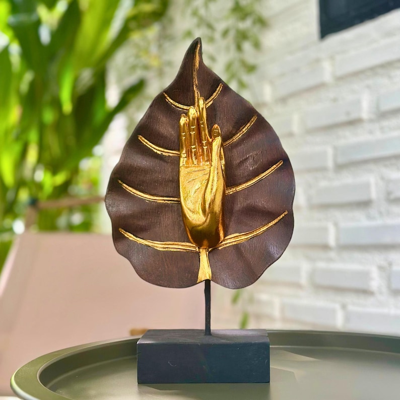 Pu&ograve; includere: Una scultura decorativa raffigurante una foglia marrone con dettagli dorati e una mano dorata. La scultura &egrave; montata su una base e uno stelo neri. La foglia ha venature dorate e una mano dorata al centro.