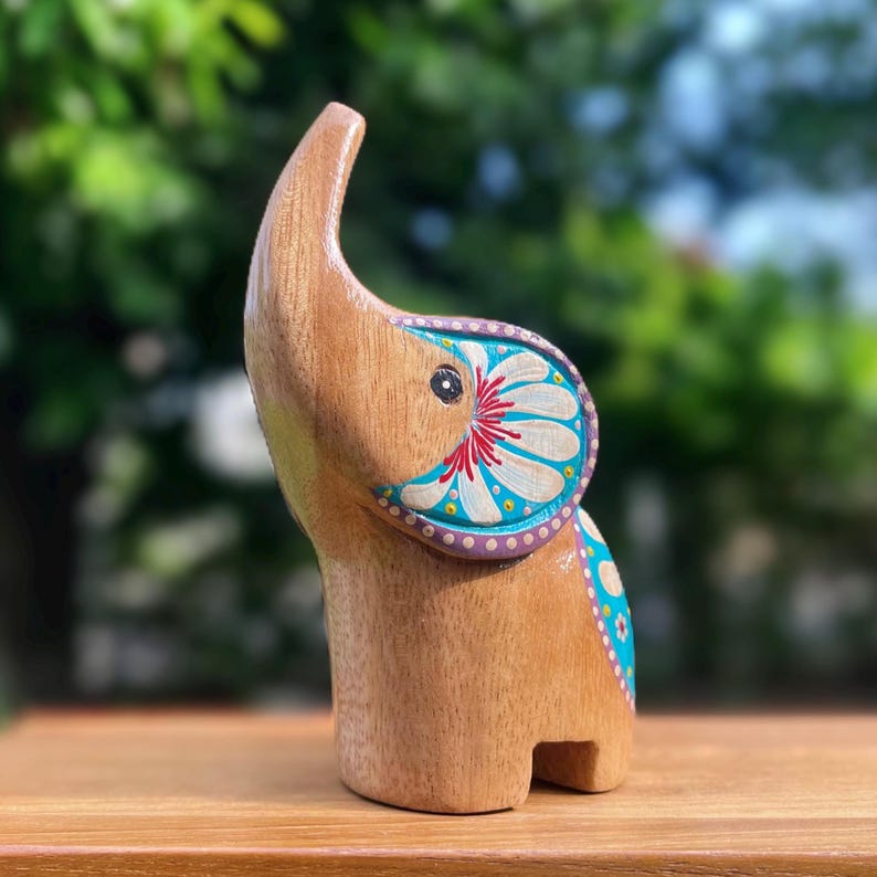 May include: Wooden elephant figurine with a trunk raised upwards. The elephant is painted with a floral design in white, red, and blue on its ear and side. The figurine is made of light brown wood and stands on a wooden surface.