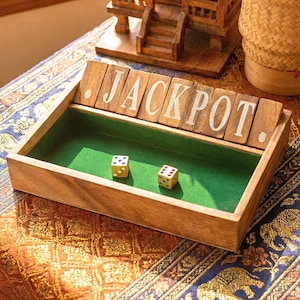 Può includere: Gioco di dadi in legno con la scritta "JACKPOT" in lettere bianche. Due dadi con puntini bianchi sono all'interno del vassoio foderato di feltro verde. Il gioco è appoggiato su una superficie a fantasia.