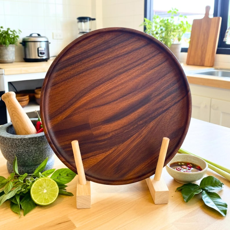 May include: A large, round, dark brown wooden serving tray with a natural wood grain pattern. The tray is displayed on a wooden stand. Kitchen items like a mortar and pestle, lime, and herbs are in the foreground.