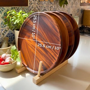 May include: A stack of four round wooden plates, each 10 inches in diameter, displayed in a wooden plate rack. The plates have a rich, dark brown wood grain pattern. The rack is made of light-colored wood.