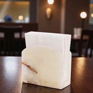 May include: A rectangular napkin holder made of translucent, cream-colored stone with a brown vein. The holder has a curved front and holds a stack of white paper napkins. The holder sits on a dark wooden table.