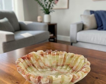May include: A shallow, round bowl crafted from polished stone featuring a scalloped, ruffled edge. The material displays a natural, translucent banded pattern with swirling veins of pale green, cream, and reddish-brown colors throughout the surface.