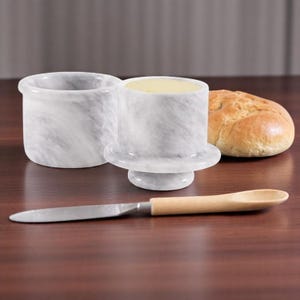 May include: White marble butter dish with a lid and a small knife. The dish is on a wooden table with a roll of bread.