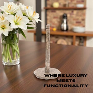 May include: A stone paper towel holder with a cylindrical post and a round top. The base is a gray, multi-sided shape. The text "WHERE LUXURY MEETS FUNCTIONALITY" is at the bottom of the image. A vase of white flowers is in the background.