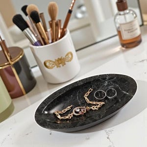 May include: A black marble oval tray holds a gold bracelet with a gemstone, and two silver rings. Makeup brushes are in a white ceramic holder with a gold design. A perfume bottle is in the background.