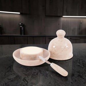 May include: A light beige marble butter dish with a matching dome-shaped lid and a small butter knife featuring a coordinating marble handle. The dish holds a round block of yellow butter, showcasing the natural veining of the polished stone.