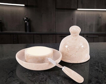May include: A light beige marble butter dish with a matching dome-shaped lid and a small butter knife featuring a coordinating marble handle. The dish holds a round block of yellow butter, showcasing the natural veining of the polished stone.