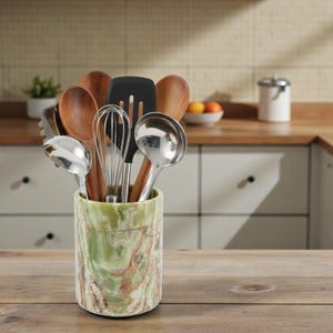 May include: A kitchen utensil holder made of green and brown marble-like material, filled with various cooking tools. The utensils include wooden spoons, a whisk, a black spatula, and a metal ladle. The holder sits on a wooden surface.