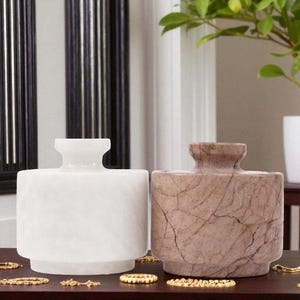 May include: Two decorative stone vases. One is white, the other is brown with a marbled pattern. Both have a cylindrical body and a small, flared neck. The vases are displayed on a dark wooden surface.