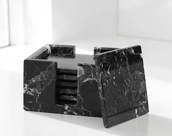 May include: A set of square black marble coasters with white veining, housed in a matching black marble holder. The set includes several stacked coasters, with one coaster positioned upright against the front of the open-sided storage box.