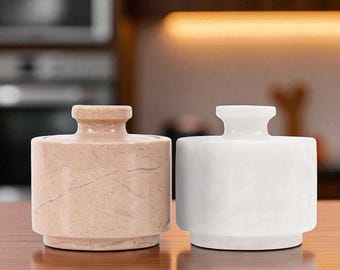 May include: Two stone jars with lids. One jar is beige with a marbled pattern, and the other is white. Both jars have a cylindrical body and a small, round handle on the lid. They are sitting on a wooden surface.