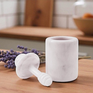 May include: A white marble mortar and pestle set. The mortar is a cylindrical shape, and the pestle has a mushroom-like head with a ribbed handle. The set is on a wooden surface, with a sprig of lavender in the background.