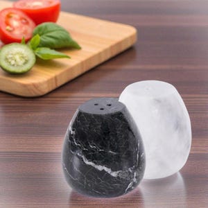 May include: A pair of marble salt and pepper shakers. One shaker is black with white veining, and the other is white. Each shaker has a rounded, egg-like shape with three holes on top for dispensing. The shakers are on a wooden surface.