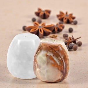 May include: Two decorative salt and pepper shakers. One is white, the other is a mix of brown, beige, and white. The shakers are egg-shaped with small holes on top. Star anise and peppercorns are in the background.
