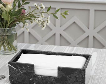 May include: A rectangular black marble tissue box with a cut-out on the front. The tissue box is filled with white tissues. A vase of flowers is in the background.