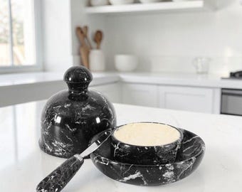 May include: A black marble butter keeper with a matching dome lid and a small spreader knife. The butter keeper features white veining throughout the stone surface. The set includes a circular base, a rounded lid with a knob handle, and a spreader.