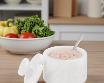 May include: A white marble salt cellar with a lid and a small stainless steel spoon. The container is filled with pink Himalayan salt. A bowl of fresh vegetables is in the background.