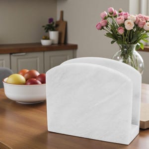 May include: A white marble napkin holder with a curved top. The holder is rectangular and sits on a wooden table. A bowl of fruit and a vase of pink roses are in the background. The napkin holder is a kitchen or dining room accessory.
