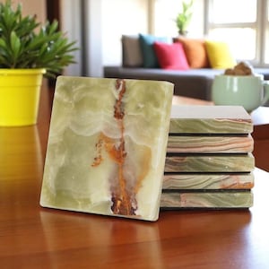 May include: A stack of six square coasters made of green onyx stone. The coasters have a natural, layered appearance with brown and white veining. One coaster is standing upright, showcasing the stone's texture and color variations.