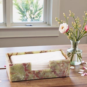 May include: A rectangular tissue box made of green and red marble. The box is filled with white tissues and sits on a wooden table. A vase of flowers is in the background.