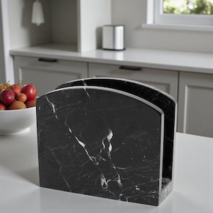 May include: A black marble napkin holder with white veining. The holder has a rectangular base and a curved top, designed to hold napkins. The kitchen accessory is made of a solid, heavy material.