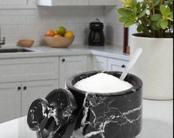 May include: A black marble sugar bowl with a lid, filled with white sugar, and a small spoon. The bowl has a circular shape with white veining. The lid is off to the side. The background shows a kitchen setting.