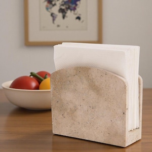 May include: A beige stone napkin holder containing white napkins. The holder has a curved top and a rectangular base. A bowl of tomatoes and a yellow pepper are in the background.