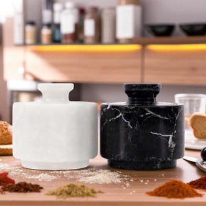 May include: Two marble spice jars, one white and one black with white veining, sit on a wooden surface. Various spices are scattered around the jars. The jars have a cylindrical shape with a small, rounded top.