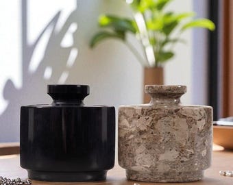 May include: Two decorative vases. One is solid black, the other is a beige and brown marbled stone. Both have a cylindrical body and a small, rounded neck. The vases are on a wooden surface with jewelry.