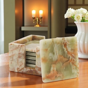 May include: A set of square coasters made of green and tan marble-like stone. The coasters are in a matching holder. The holder and coasters have a natural, veined pattern. A white vase with white flowers is in the background.
