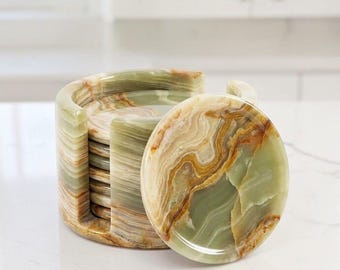 May include: A set of circular stone coasters featuring natural bands of sage green, cream, and amber brown. The coasters are stacked vertically within a matching cylindrical stone holder that has a side opening for easy access to each individual piece.