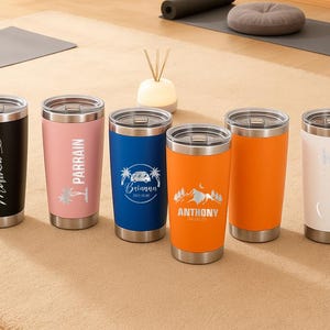 May include: Six stainless steel insulated tumblers in black, pink, blue, orange, and white. Each tumbler has a silver lid and base. The tumblers are personalised with names and graphics, including a heart, palm trees, a camper van, and mountain scene.