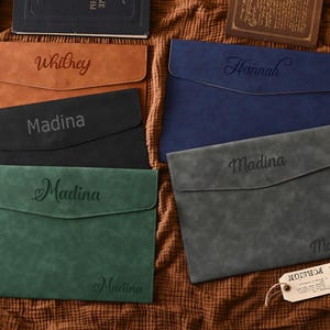May include: A selection of personalised leather document holders in shades of brown, black, green, blue, and grey. Each holder has a name engraved on the front. The holders are displayed on a textured brown surface.