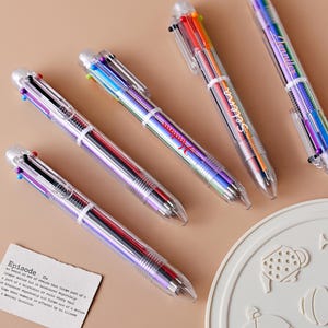 May include: Four multi-coloured pens with clear plastic barrels. Each pen has six different coloured ink refills. The pens are arranged in a row on a light brown surface.