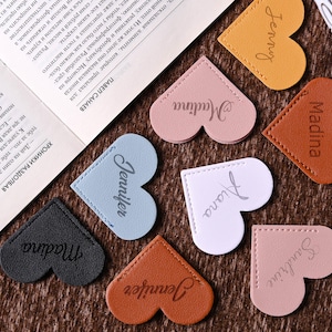 May include: Heart-shaped bookmarks in various colors, including tan, pink, and blue, are displayed. Each bookmark has a name printed on it, such as "Jenny" and "Madina". The bookmarks are made of a leather-like material and are placed near an open book.