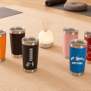 Puede incluir: Una colecci&oacute;n de vasos aislados en varios colores, incluyendo naranja, negro, rosa, azul y rojo. Cada vaso tiene una tapa plateada y texto o gr&aacute;ficos personalizados. Los vasos se exhiben sobre una alfombra de tono neutro.