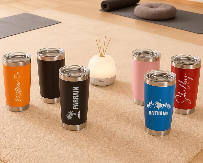 Puede incluir: Una colecci&oacute;n de vasos aislados en varios colores, incluyendo naranja, negro, rosa, azul y rojo. Cada vaso tiene una tapa plateada y texto o gr&aacute;ficos personalizados. Los vasos se exhiben sobre una alfombra de tono neutro.